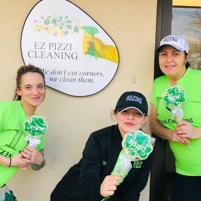 Three women in green shirts standing in front of a pizza cleaning sign.